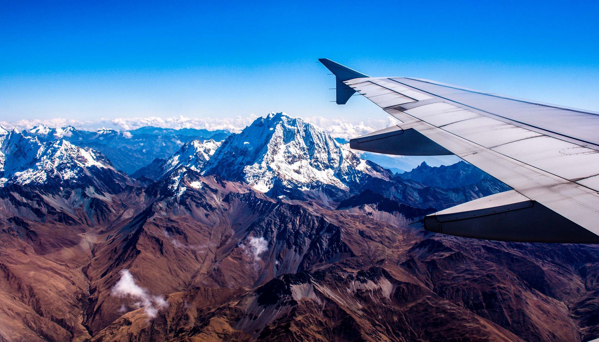 View from an airplane window showing a snowy mountain range under a clear blue sky, with the plane’s wing in the foreground—reminding travelers to always review booking conditions before departure.