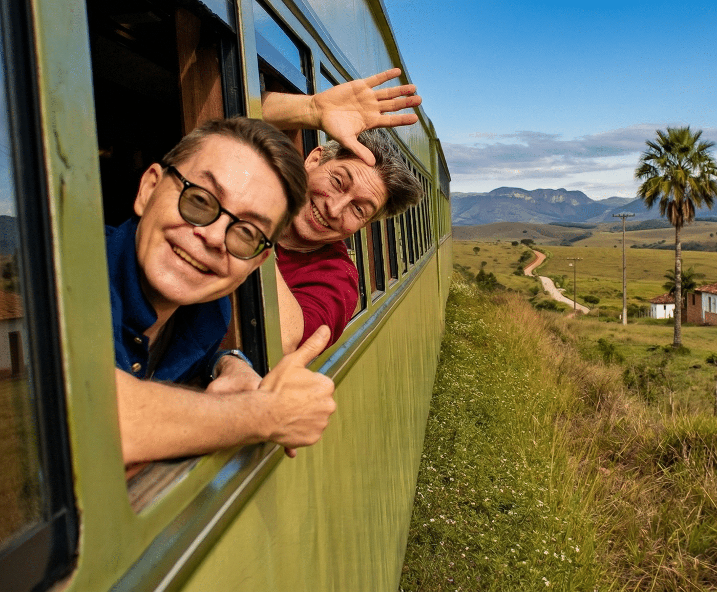 Two smiling people lean out of the windows of a green train—one giving a thumbs up, the other waving—showcasing one of the top 10 great reasons to travel, with grassy fields, a palm tree, and mountains under a blue sky in the background.