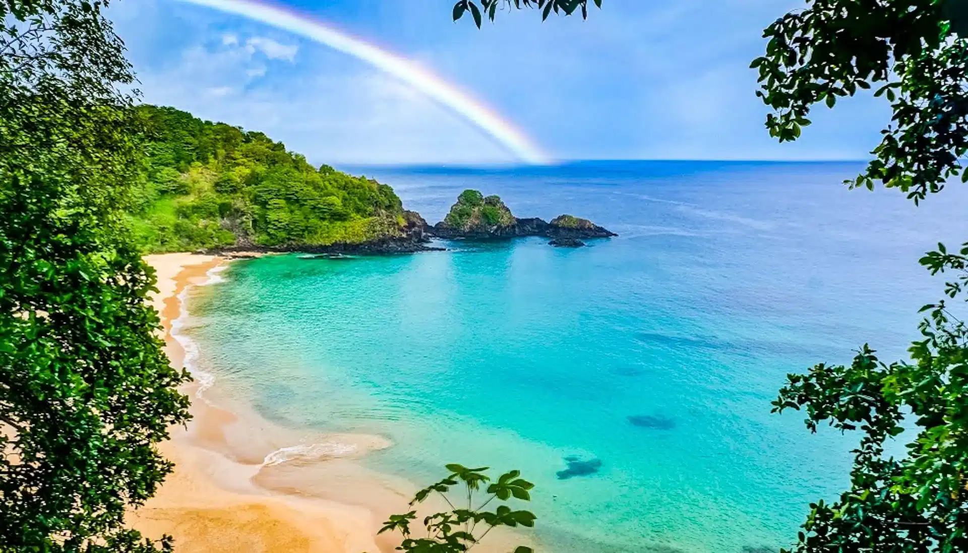 A tropical South America beach with golden sand and clear turquoise water, surrounded by green foliage. A rainbow arcs across the blue sky above rocky islets—experience this paradise with our exclusive South America Specials travel deals.