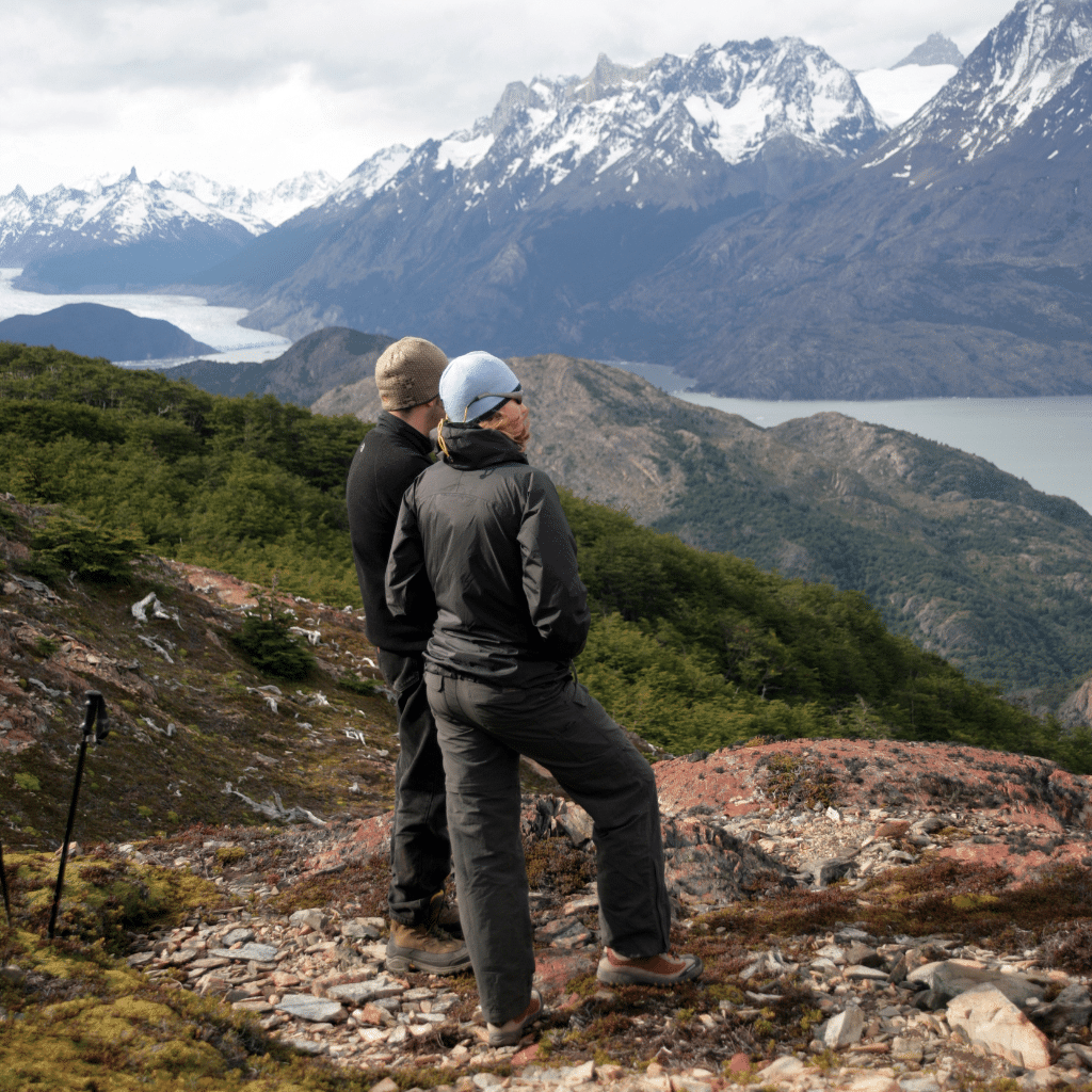 Two people wearing jackets and hats stand on a rocky hillside, overlooking a scenic view of mountains, green valleys, and a winding river under a cloudy sky—book with confidence to explore breathtaking landscapes like this.