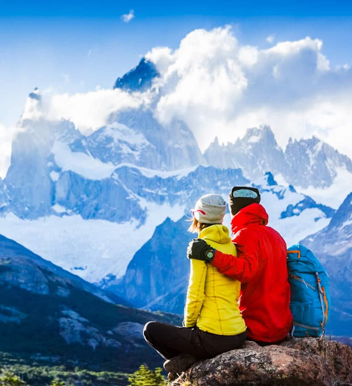 A couple wearing bright jackets and winter hats sits on a rock, embracing while admiring a dramatic snow-covered mountain landscape—an ideal scene to reflect on how custom planning works for your next adventure.