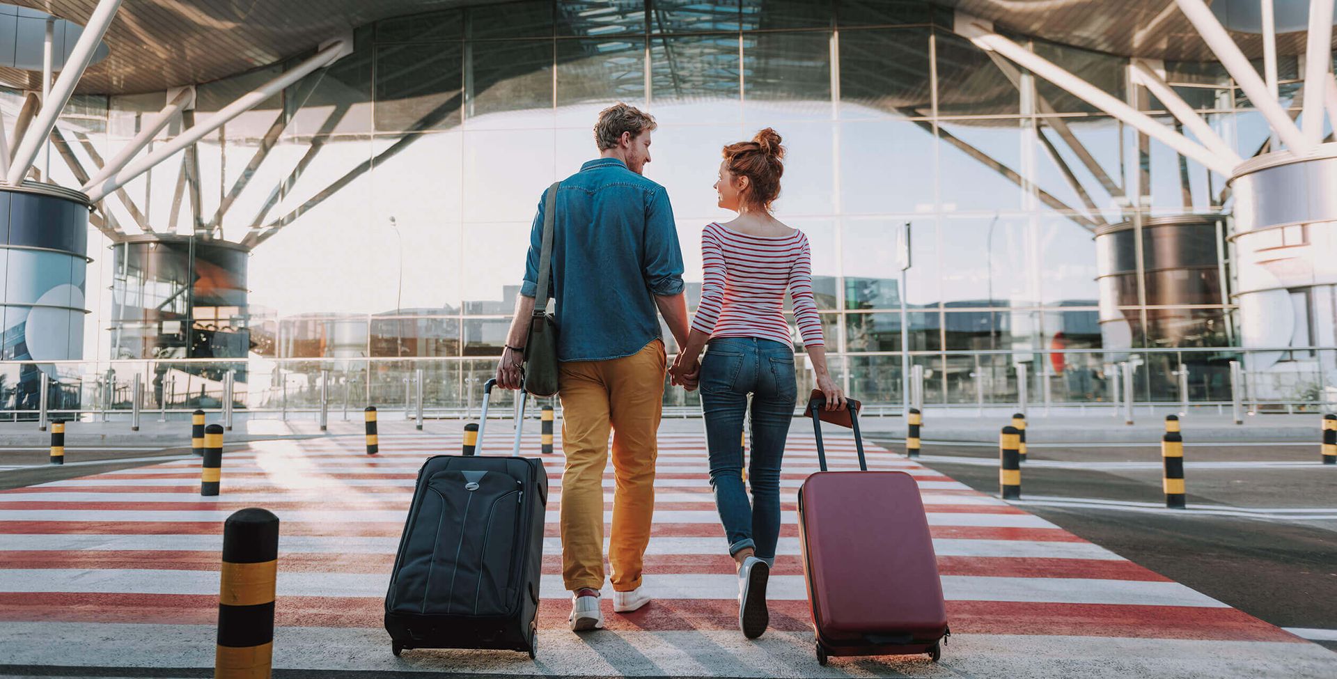 A couple holding hands, walking across a crosswalk toward a modern airport terminal, each pulling a suitcase. The scene suggests travel or departure.