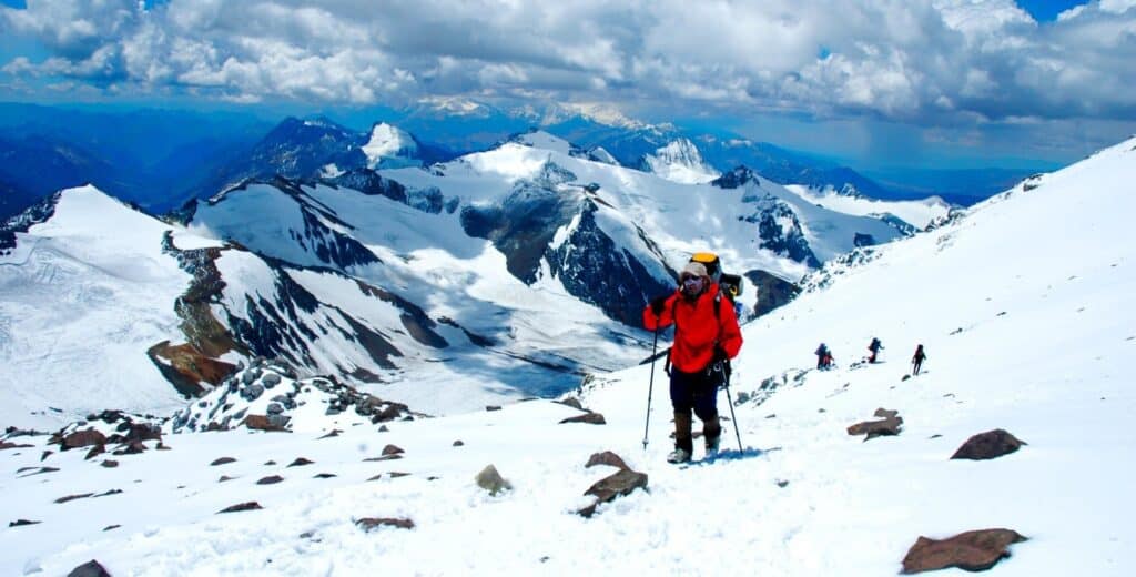A group of climbers in winter gear ascends a snow-covered mountain slope, surrounded by rugged, snow-capped peaks under a partly cloudy sky—a scene echoing the history of great SouthAmerica.travel expeditions. The lead climber is wearing red and carrying a backpack.