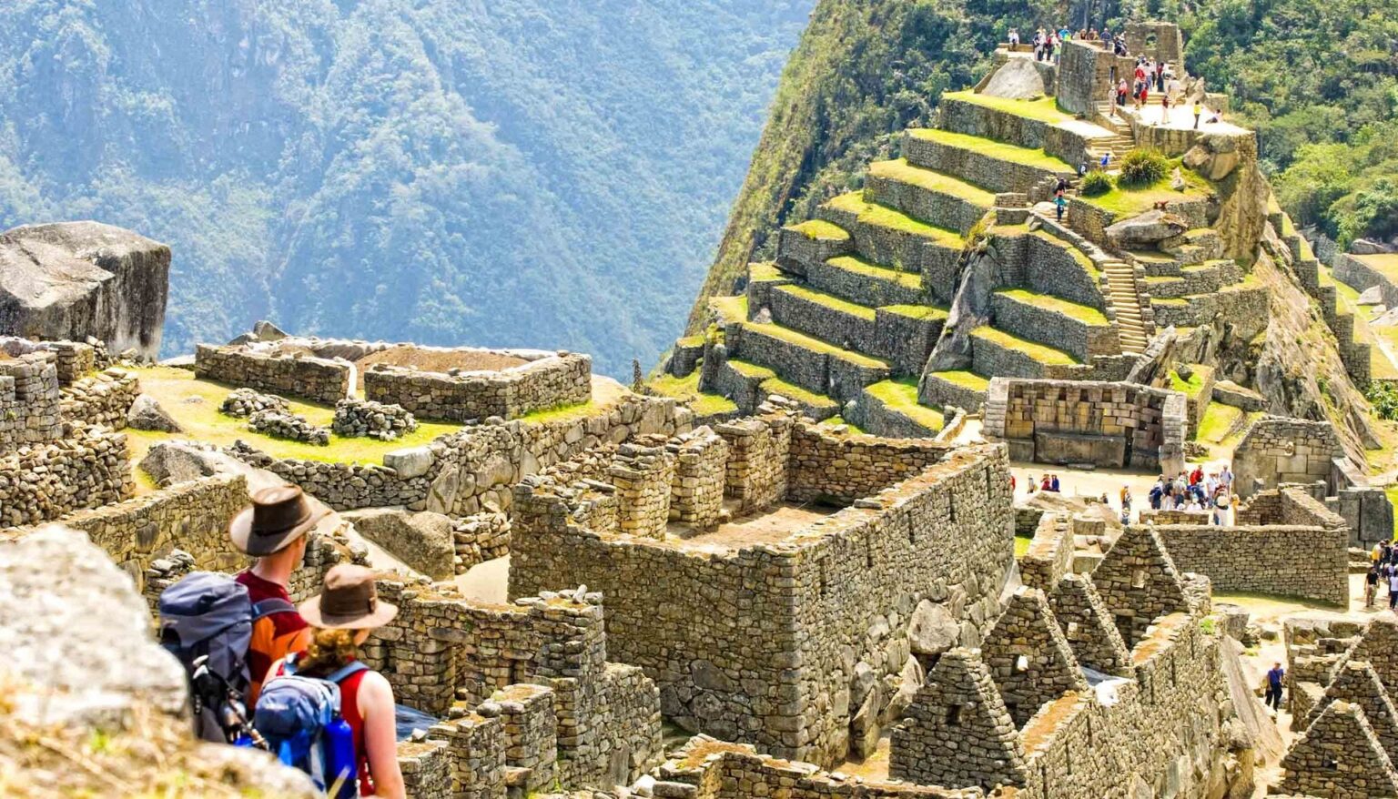 Tourists with backpacks and hats explore the ancient stone ruins and terraced hillsides of Machu Picchu, surrounded by lush mountains and bright daylight—sign up for our newsletter to discover more unforgettable travel destinations.