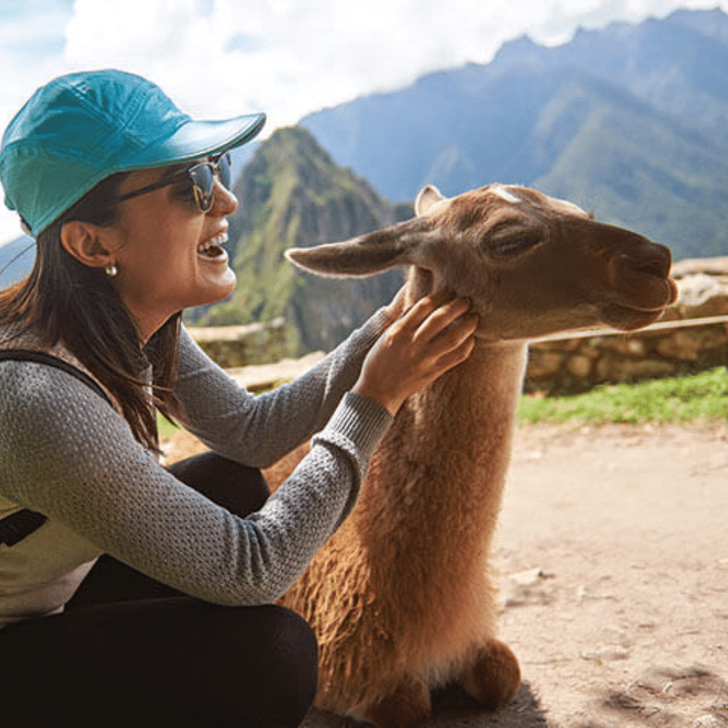 A smiling woman in a turquoise cap pets a llama while crouching outdoors, with green mountains and ancient stone ruins in the background—an adventure you can book with confidence.