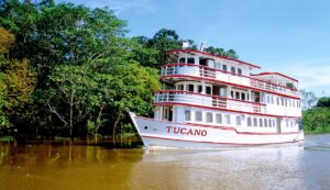 A white and red riverboat named Tucano cruises along a brown, calm river, passing lush green trees under a clear blue sky.
