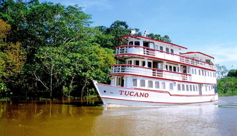 A white and red riverboat named Tucano cruises along a brown, calm river, passing lush green trees under a clear blue sky.