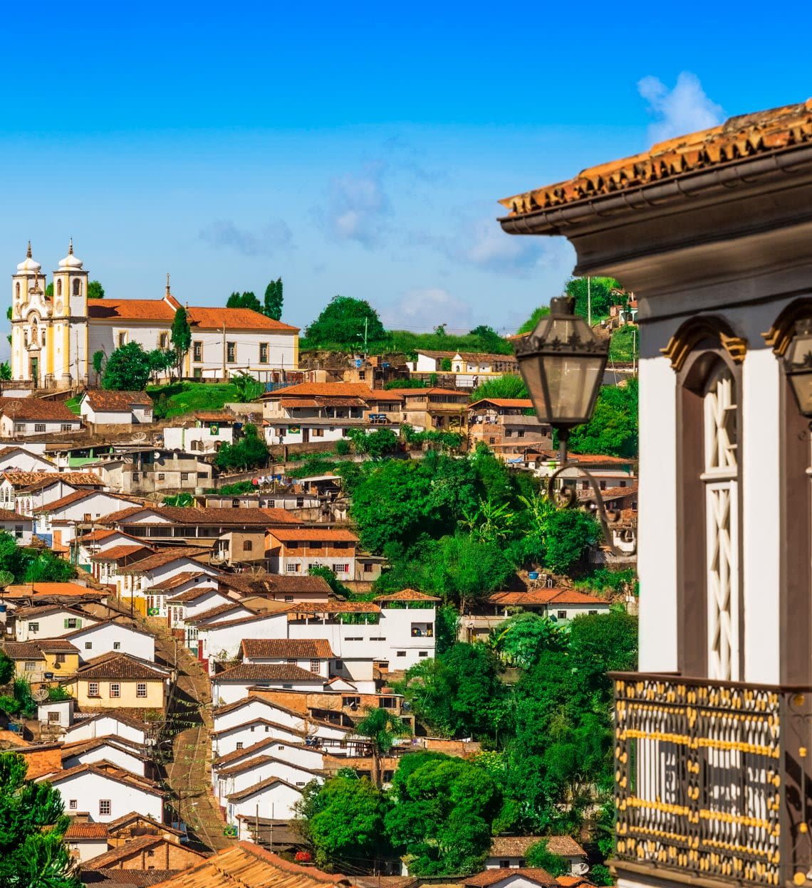 View of a historic town with red-tiled roofs, white colonial buildings, and a church on a hill—one of the top reasons to visit. Enjoy lush greenery and a traditional balcony with lantern in the foreground under a bright blue sky.