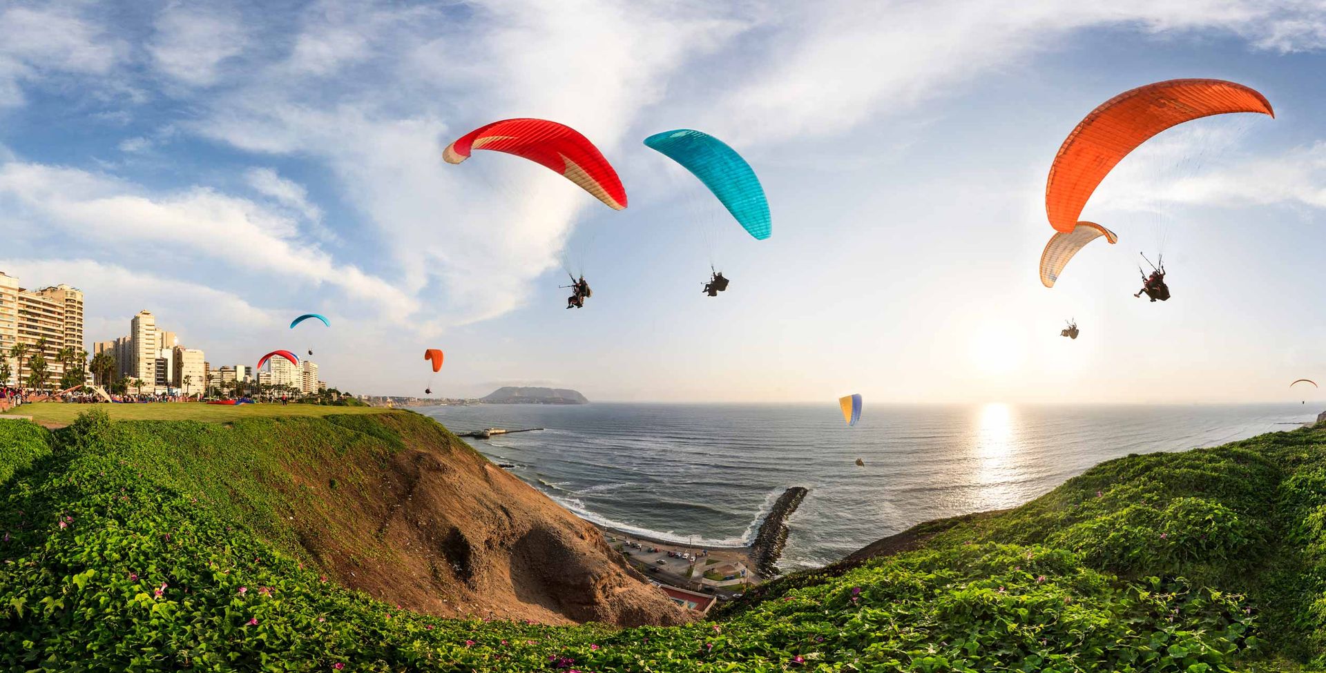 Paragliders soar above a coastal cliff with green foliage, overlooking the ocean at sunset. Tall buildings are visible on the left, and colorful parachutes fill the sky—each flight as unique as a custom planning process in motion.