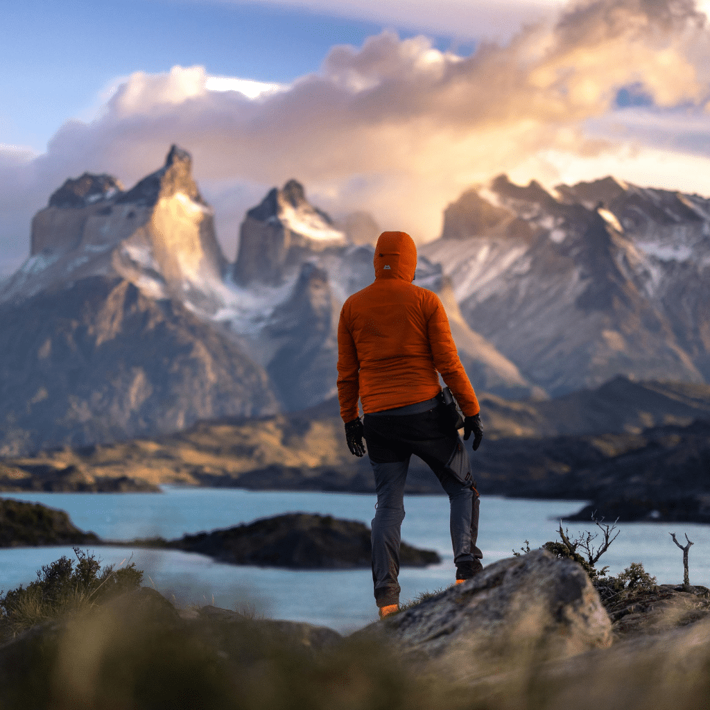 A person in an orange jacket stands on a rocky hill, overlooking a turquoise lake and dramatic, snow-capped mountains under a cloudy South America sky at sunset.