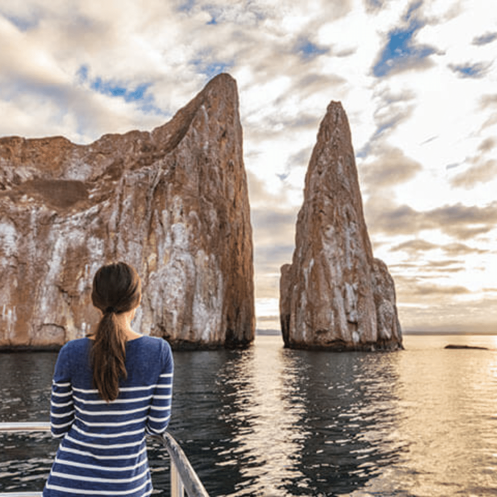 A person with dark hair in a ponytail and a blue striped sweater stands on a boat, looking at two large rock formations rising from the ocean under a partly cloudy sky—an inspiring view to book with confidence for your next adventure.