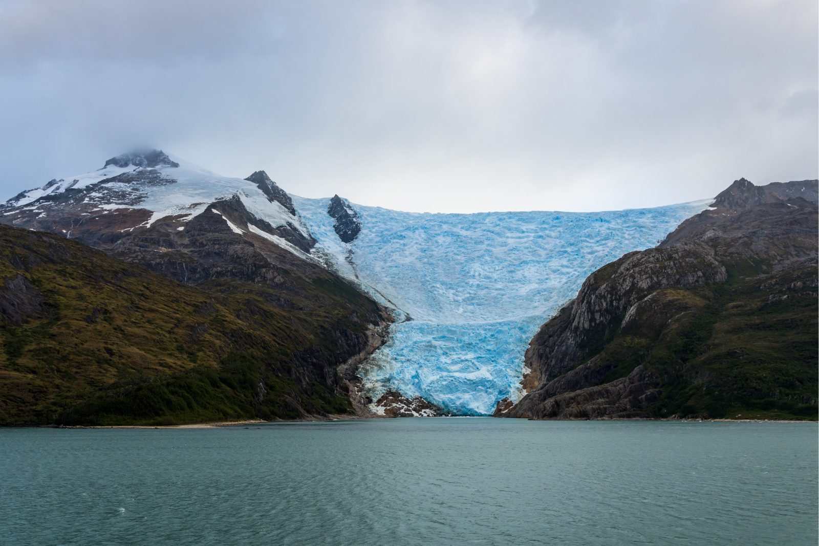 A blue tidewater glacier spilling between forested slopes in a Fuegian fjord off the Beagle Channel