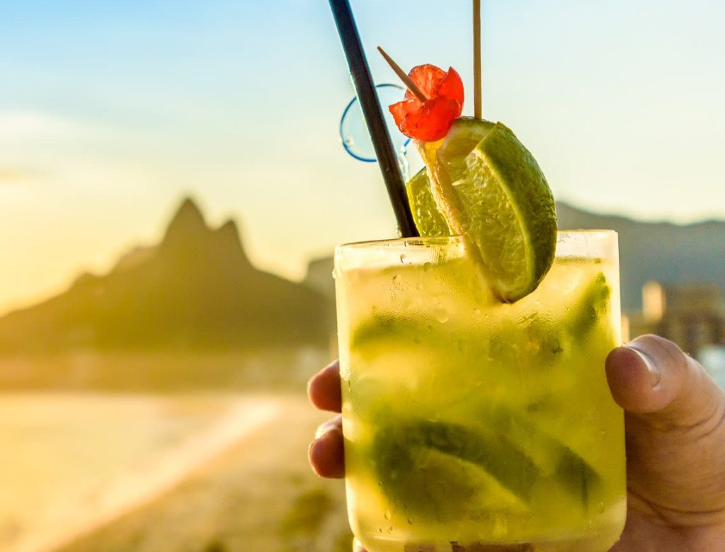 A hand holds a glass of caipirinha garnished with lime wedges, cherries, a straw, and a cocktail umbrella—one of the top reasons to enjoy sunsets by the beach and mountains in pure relaxation.