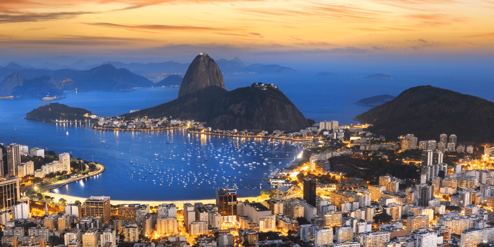 Aerial view of Rio de Janeiro at dusk, showing city buildings, Guanabara Bay with boats, and the iconic Sugarloaf Mountain in the background, under a purple-blue evening sky.