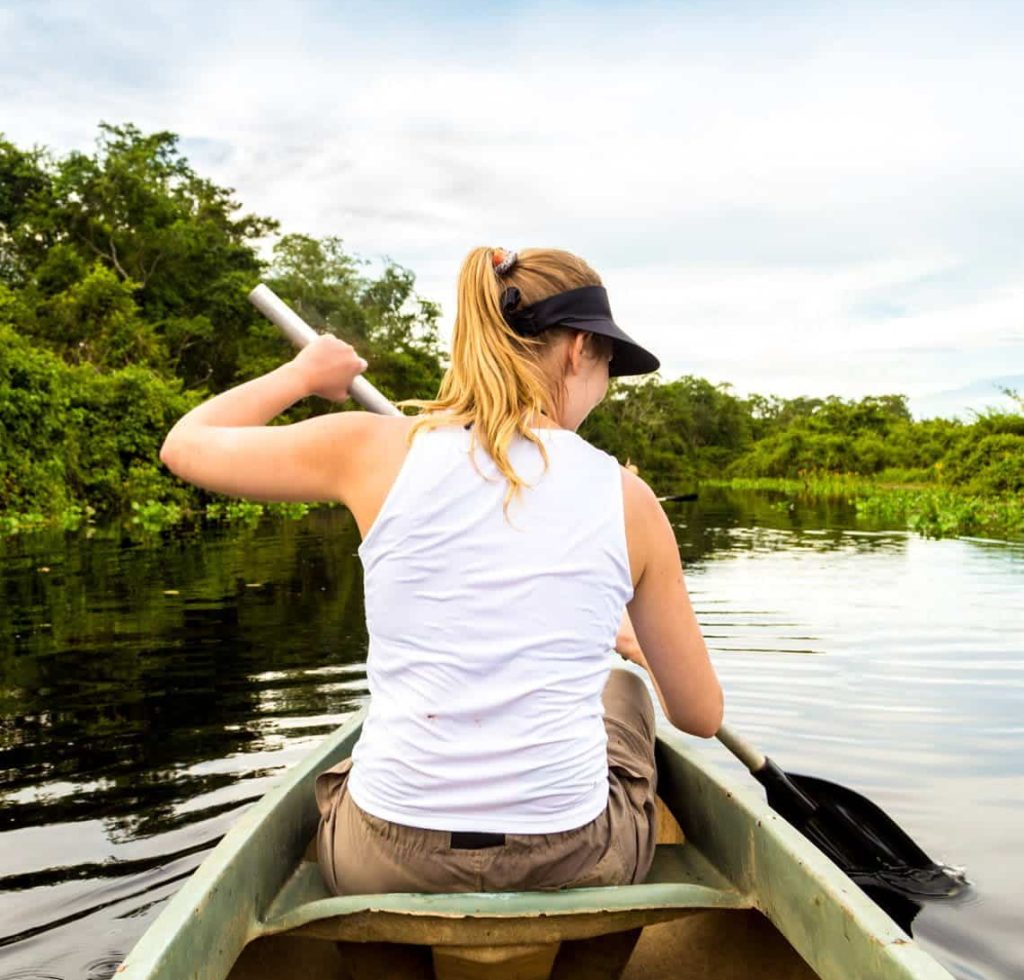 A woman with blonde hair in a ponytail paddles a canoe down a calm river, surrounded by lush green trees and a partly cloudy sky—a peaceful moment that echoes the beauty of personalized planning. She wears a white sleeveless top, visor, and sunglasses.