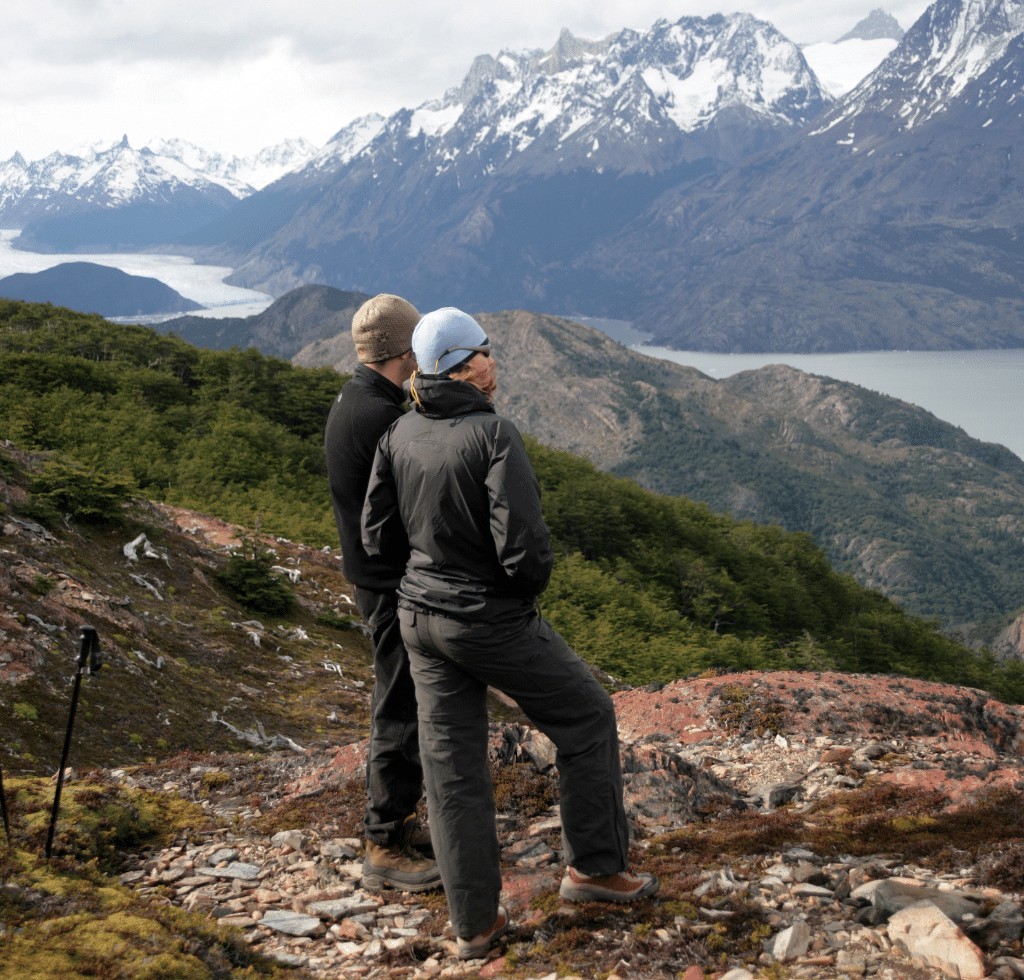 Two people wearing jackets and hats stand on a rocky hillside, overlooking a scenic view of mountains, green valleys, and a winding river under a cloudy sky—book with confidence to explore breathtaking landscapes like this.