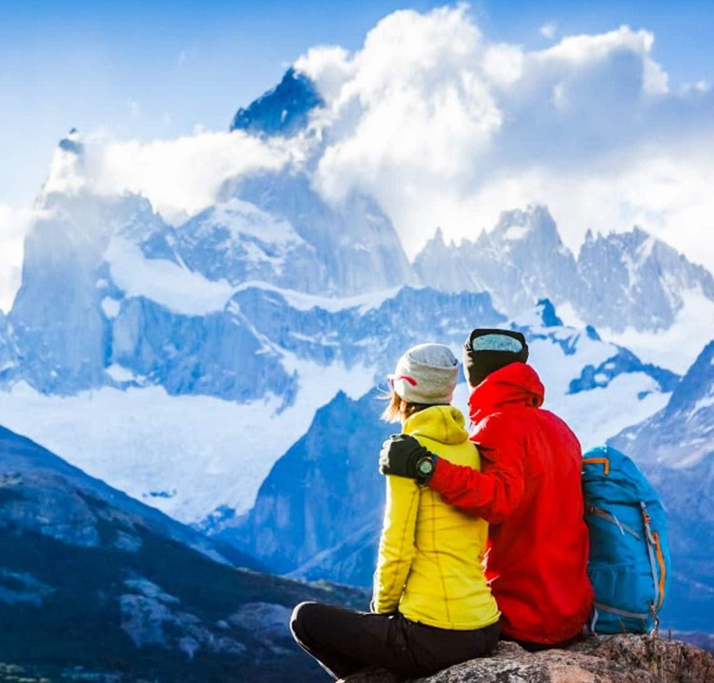 A couple wearing bright jackets and winter hats sits on a rock, embracing while admiring a dramatic snow-covered mountain landscape—an ideal scene to reflect on how custom planning works for your next adventure.