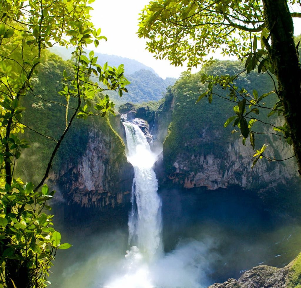 A tall, powerful waterfall cascades down a rocky cliff surrounded by lush green trees and mist—one of the top reasons nature lovers are drawn to this breathtaking spot, with majestic mountains in the background under a bright, partly cloudy sky.