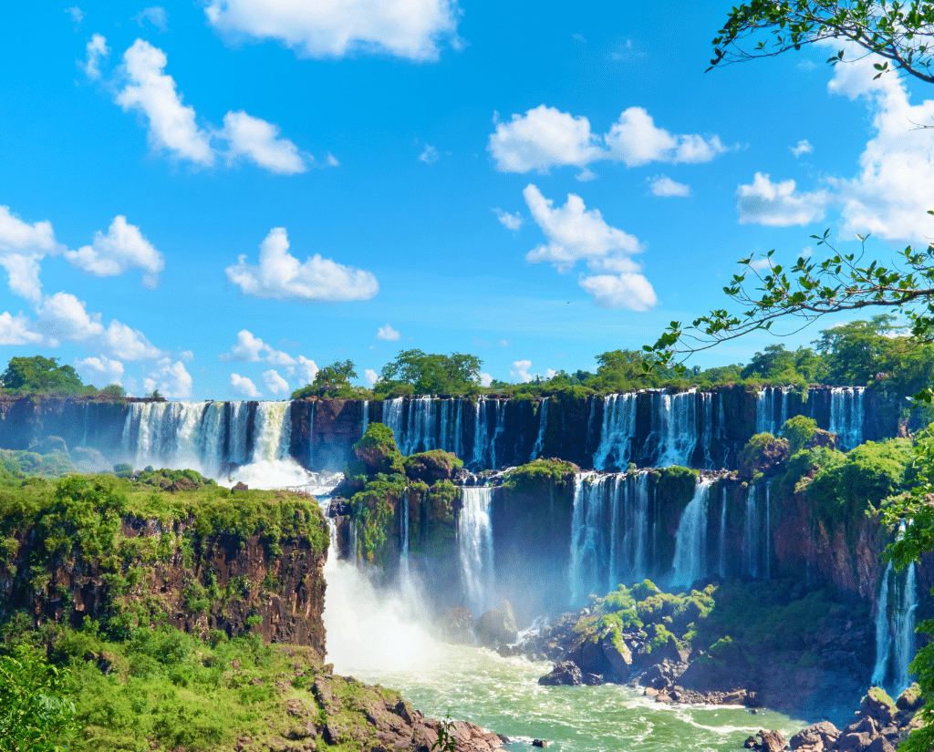 A wide view of Iguazu Falls cascading over lush green cliffs in South America, with mist rising and a bright blue sky dotted with white clouds. Dense foliage and rocky terrain surround these powerful waterfalls—perfect for South America Specials.