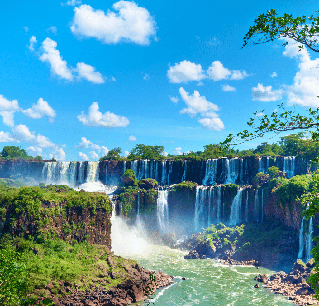 A wide view of Iguazu Falls cascading over lush green cliffs in South America, with mist rising and a bright blue sky dotted with white clouds. Dense foliage and rocky terrain surround these powerful waterfalls—perfect for South America Specials.
