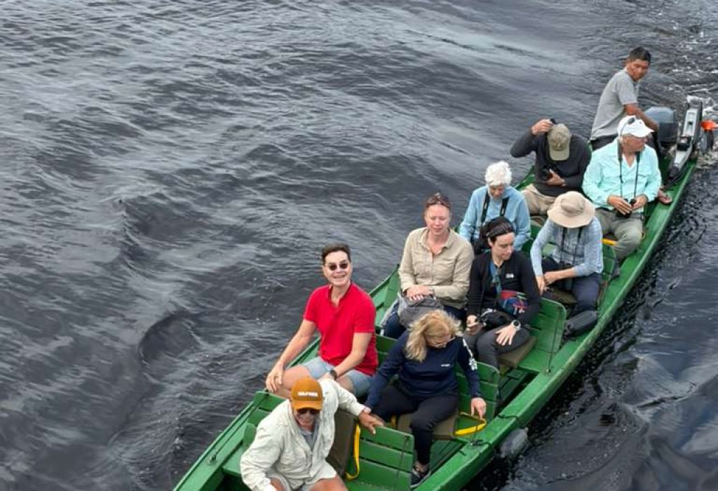A group of people sit in a green motorized boat on dark, calm water—some wearing hats and sunglasses—looking up and smiling at the camera, showing one of the top reasons to enjoy time outdoors with friends.