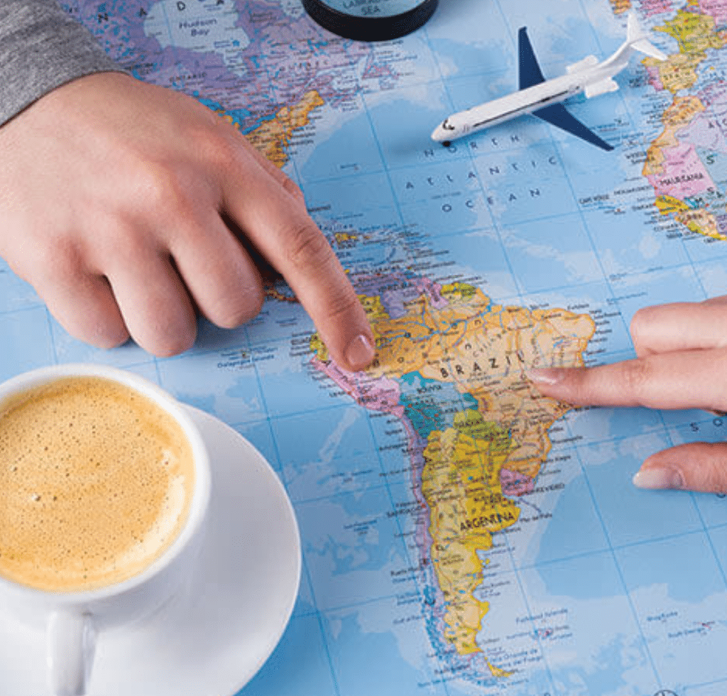 Two hands point to Brazil on a world map beside a small toy airplane. A cup of coffee sits nearby, inspiring you to book with confidence as you plan your adventure to South America.