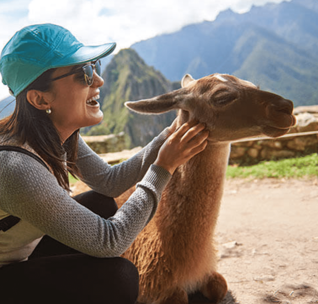 A smiling woman in a turquoise cap pets a llama while crouching outdoors, with green mountains and ancient stone ruins in the background—an adventure you can book with confidence.