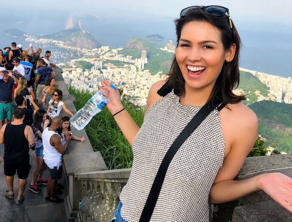 A smiling woman holds a water bottle and poses at a scenic viewpoint overlooking Rio de Janeiro—one of the 10 Great Reasons to visit—with Sugarloaf Mountain, city buildings, and other tourists visible in the background.