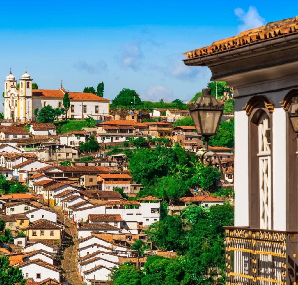 View of a historic town with red-tiled roofs, white colonial buildings, and a church on a hill—one of the top reasons to visit. Enjoy lush greenery and a traditional balcony with lantern in the foreground under a bright blue sky.