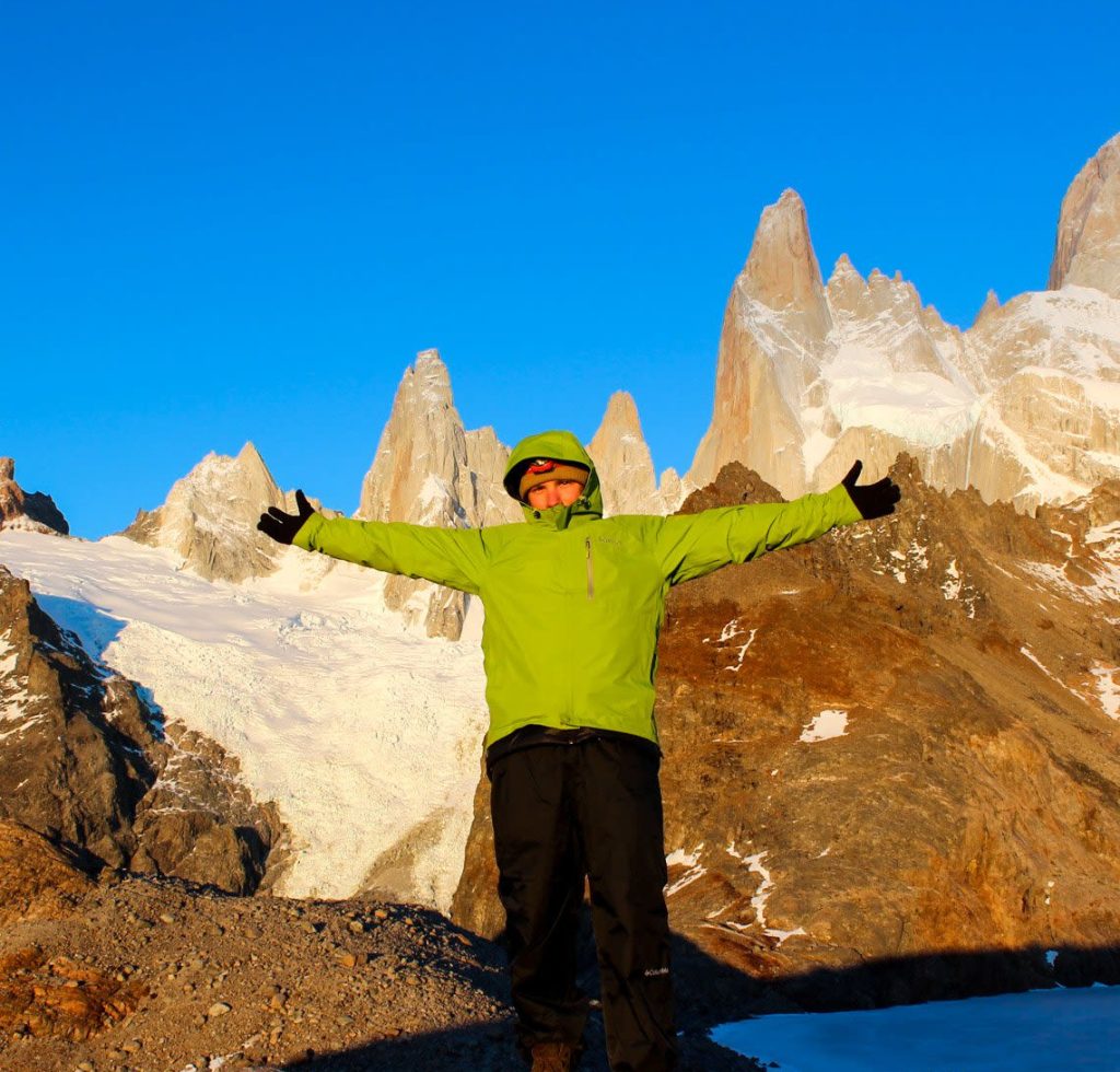 A person in a bright green jacket stands with arms outstretched on a rocky mountain, embracing the adventure—much like the freedom found in the custom planning process. Snow-covered peaks and a clear blue sky complete this breathtaking scene.