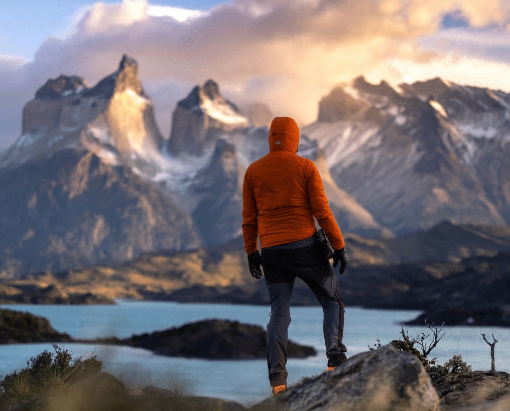 A person in an orange jacket stands on a rocky hill, overlooking a turquoise lake and dramatic, snow-capped mountains under a cloudy South America sky at sunset.