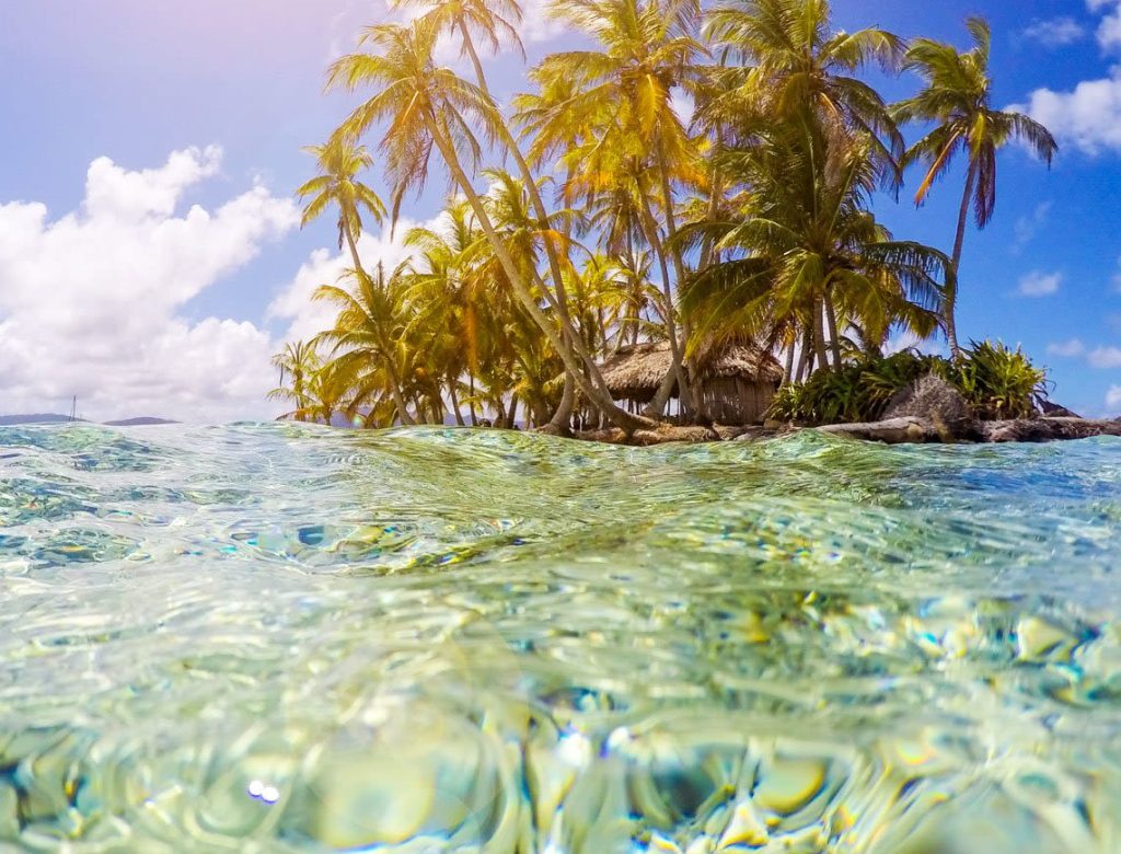 Crystal-clear turquoise water sparkles in the sunlight, with a tropical island and tall palm trees in the distance—just one of 10 great reasons to dream of this paradise under a blue sky with scattered clouds.