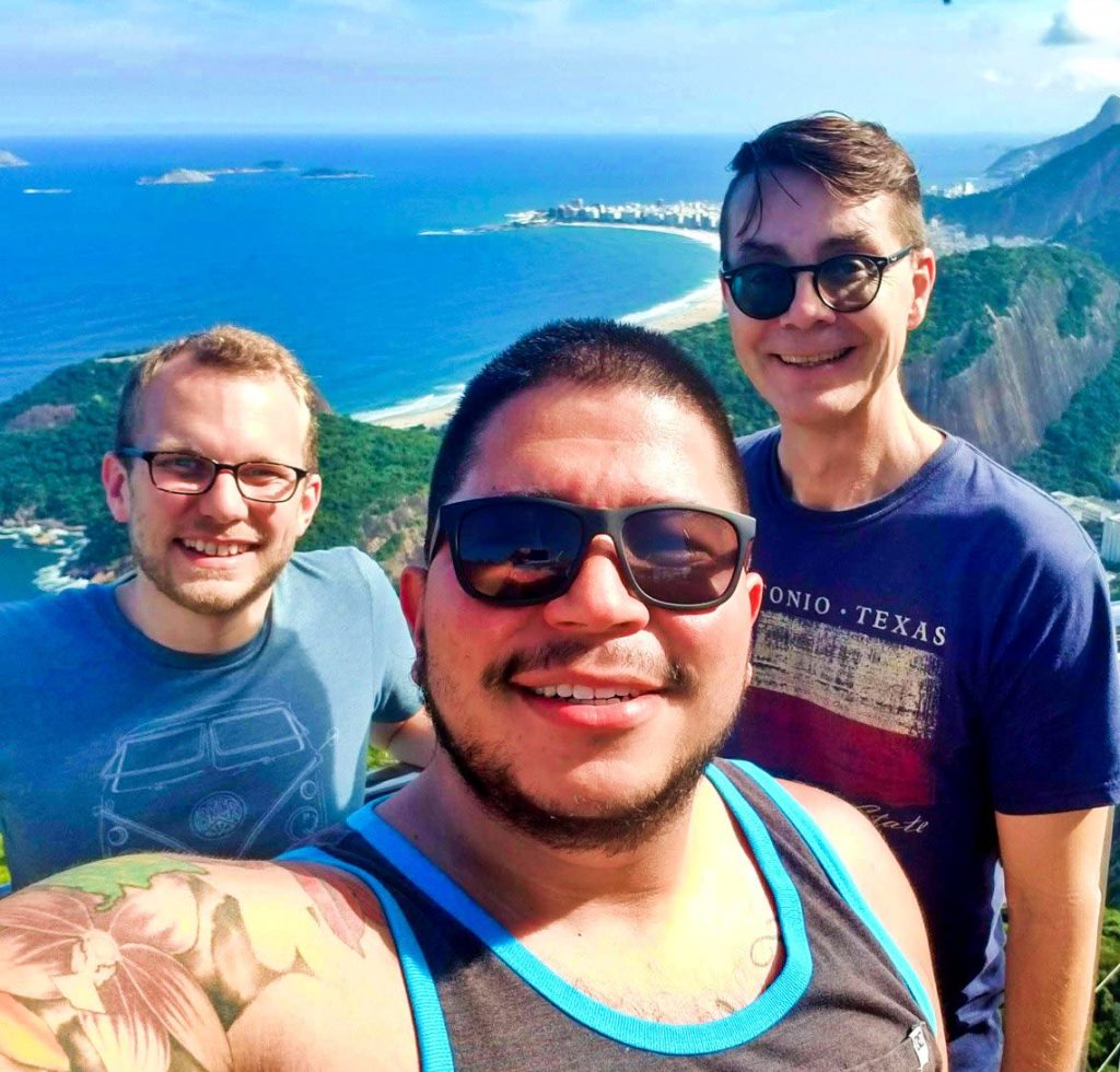 Three men smiling and posing for a selfie on a sunny day with an ocean, sandy beach, and green hills in the background. The man in front wears sunglasses and a tank top; the other two stand behind him, also smiling.