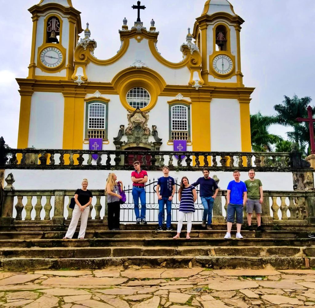 Eight people stand on stone steps in front of a yellow and white baroque church with two bell towers, arched windows, and decorative details. Palm trees and a cloudy sky are in the background.
