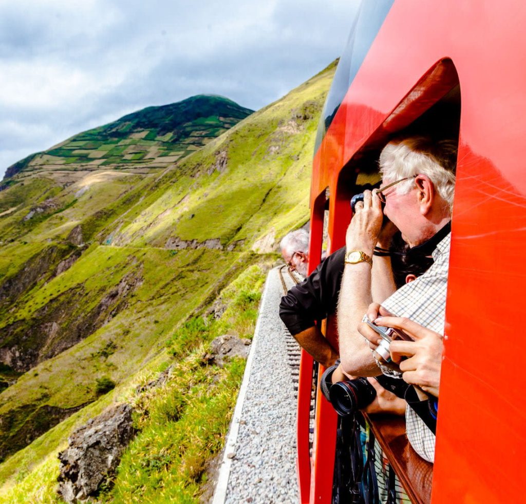 People lean out of the window of a bright red train, taking photos of green hills and mountains under a cloudy sky—a scene that offers one of the top reasons to experience this breathtaking journey along a winding track.