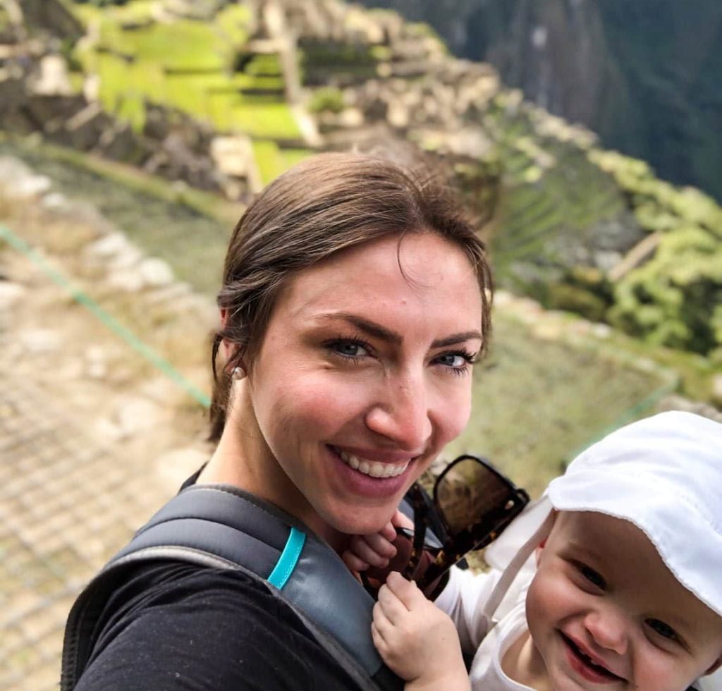A woman smiling and carrying a baby in a front carrier takes a selfie at Machu Picchu, with the ancient Incan ruins and green terraces visible in the blurred background.