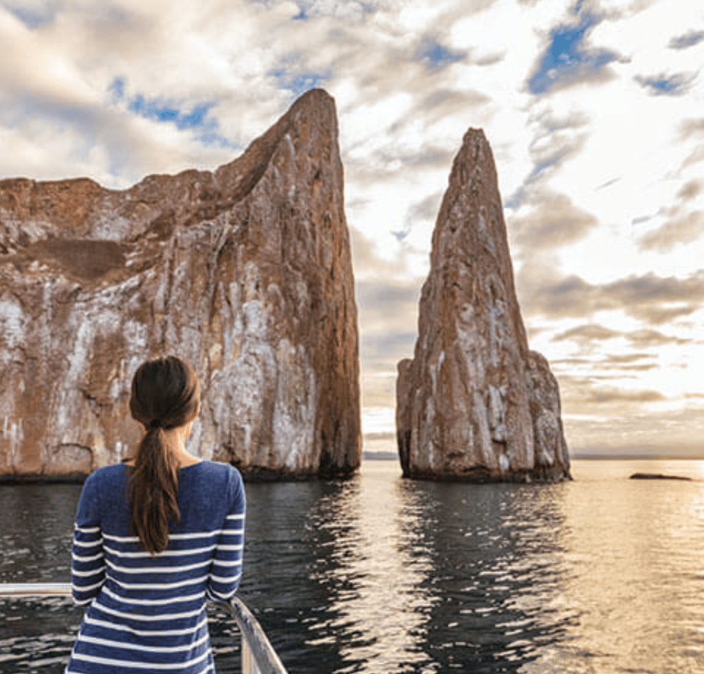 A person with dark hair in a ponytail and a blue striped sweater stands on a boat, looking at two large rock formations rising from the ocean under a partly cloudy sky—an inspiring view to book with confidence for your next adventure.