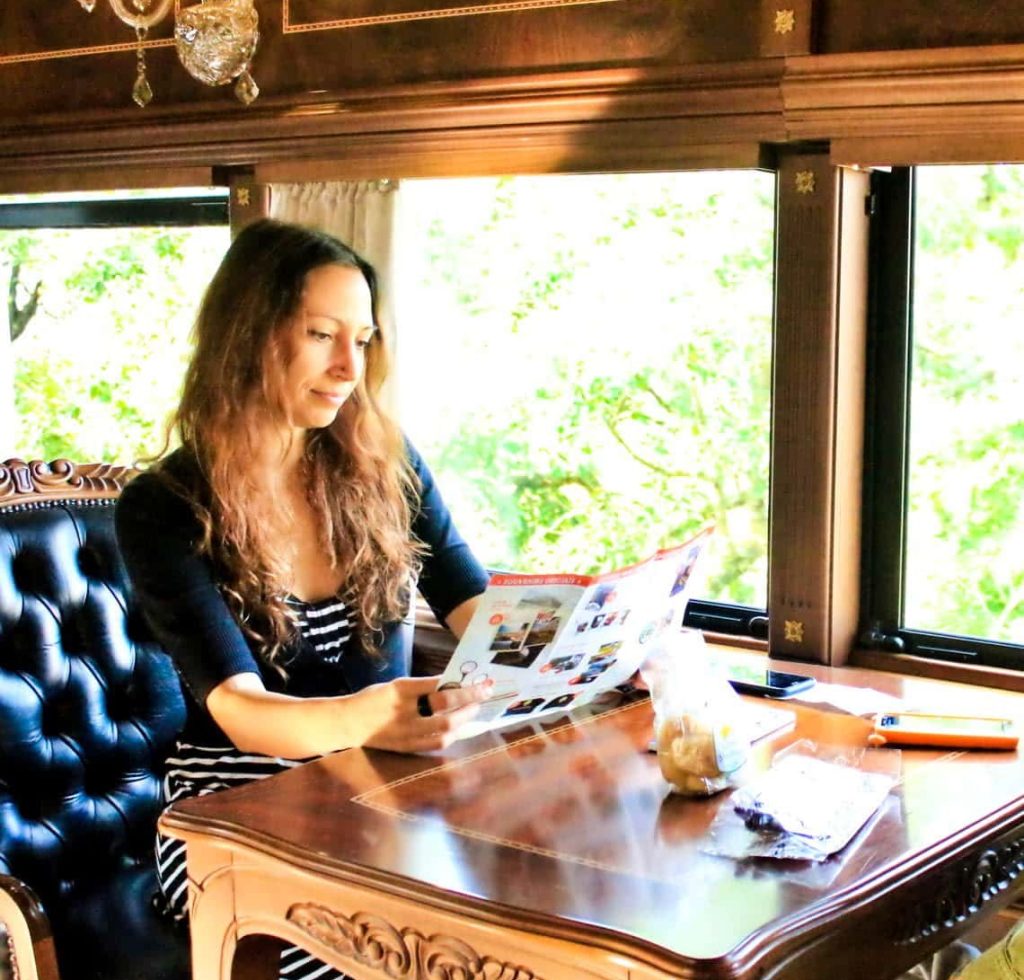 A woman with long hair sits at a polished wooden table on a vintage train, reading a colorful magazine. Sunlight streams through large windows, illuminating her and the elegant decor. Snacks and a notebook are on the table.