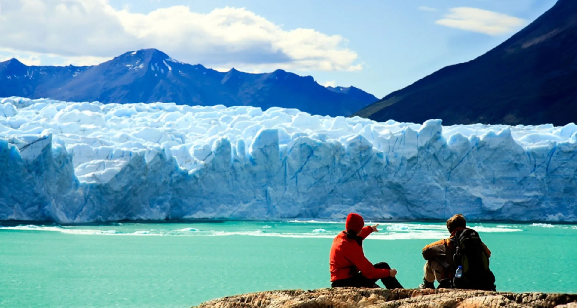 Two travelers sit to look at glacier, one pointing